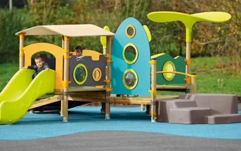Des enfants jouent sur une aire de jeux colorée, respectant la réglementation aire de jeux extérieur. Un toboggan vert vif et une structure inspirée d'une fusée bleue.