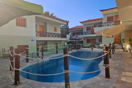 Piscine bleue entourée de poteaux en bois et de cordes, avec des bâtiments clairs et des toits rouges. Idéal pour une liste colonie de vacances.