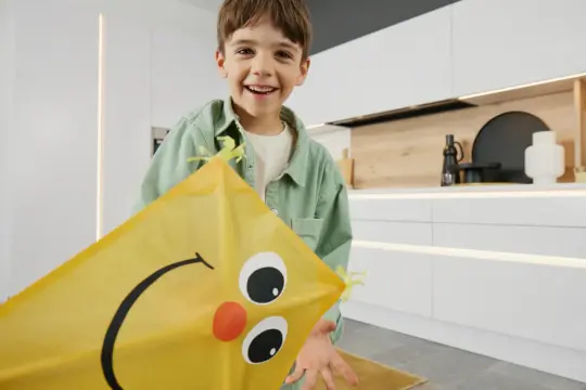 Un garçon souriant tient un cerf-volant jaune avec un visage dessiné, prêt pour une activité manuelle.