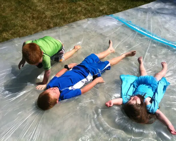 Trois enfants s'amusent sur un jeu d'eau pour kermesse, glissant sur une bâche mouillée sous le soleil.