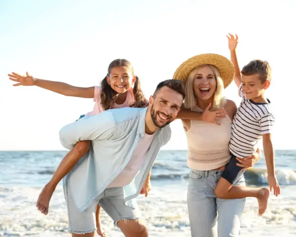 Famille souriante sur la plage, père portant sa fille sur le dos, mère tenant son fils.