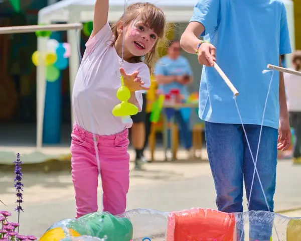 Une petite fille joyeuse participe à des jeux de kermesse pour tout petit, essayant d'attraper un jouet jaune avec une canne à pêche improvisée.