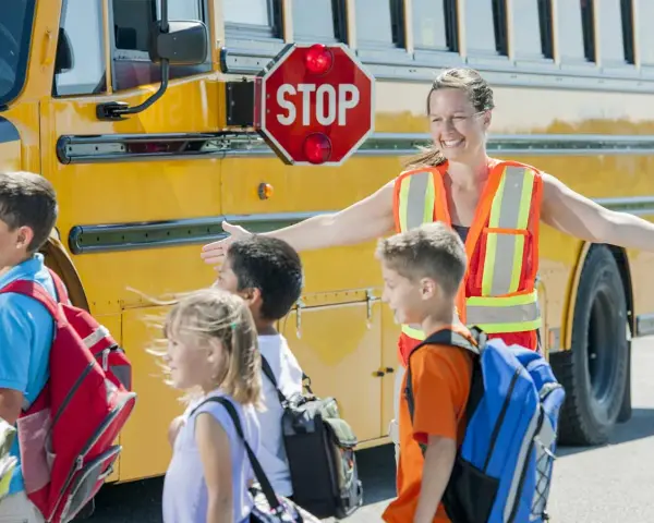 Des enfants montent dans un bus scolaire jaune, guidés par une adulte portant un gilet de sécurité orange.