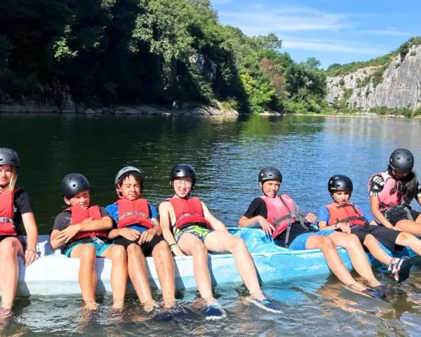 Des enfants en gilets de sauvetage et casques sur un raft bleu sur une rivière, entourés de verdure et de falaises.