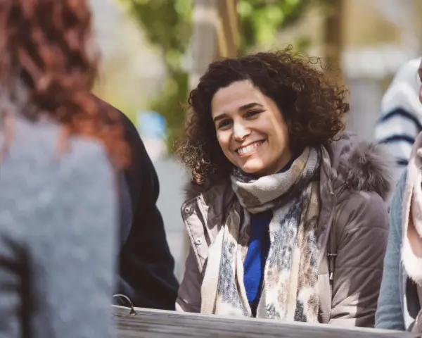 Une femme souriante aux cheveux bouclés porte une écharpe et une veste à capuche en fourrure.