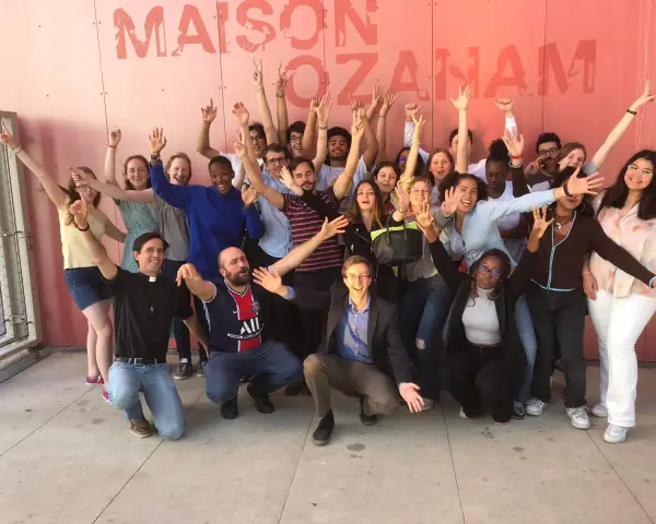 Un groupe de jeunes gens, dont un prêtre, pose joyeusement devant un mur rose avec le texte "MAISON OZANAM".