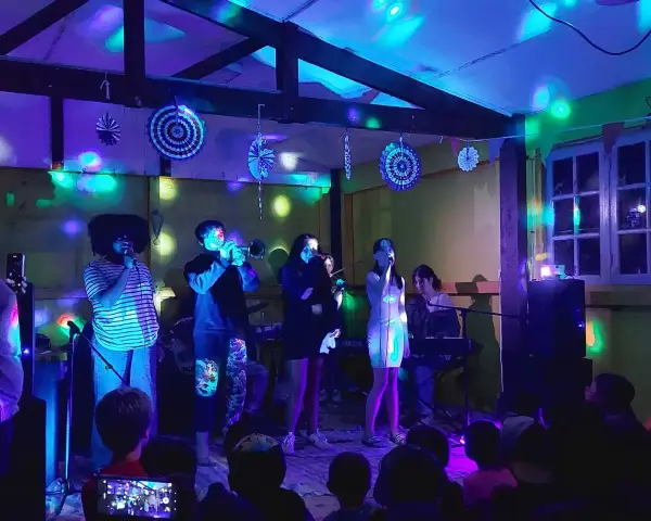Scène de concert animée avec des lumières colorées. Un groupe joue, avec des chanteurs, un trompettiste et un claviériste. Le public, dont des enfants, filme le spectacle. Ces vacances musicales sans frontières sont une fête pour les sens.