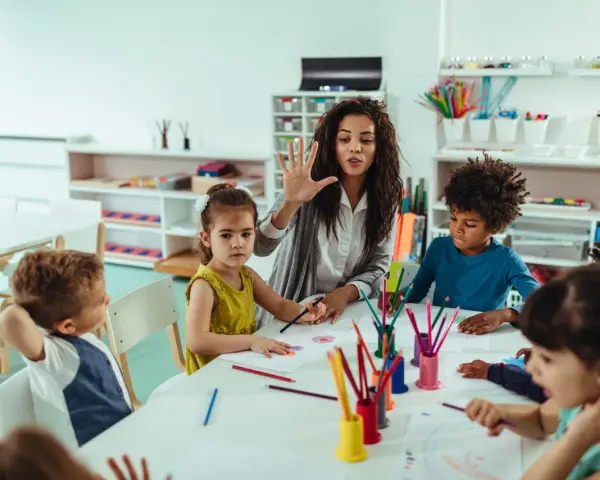 Une animatrice explique une activité créative à des enfants autour d'une table. Son rôle est d'éveiller leur curiosité.