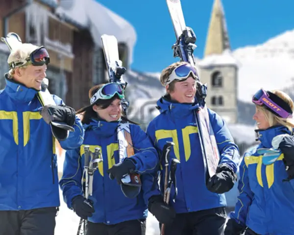Quatre jeunes skieurs souriants en tenue bleue et jaune, portant des skis et des bâtons, dans une station de ski enneigée.