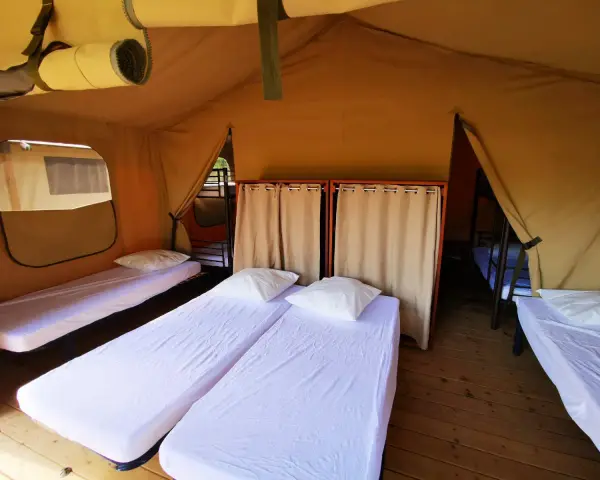 Intérieur d'une chambre de colonie de vacances avec plusieurs lits simples, draps blancs et oreillers. Des rideaux séparent les espaces.