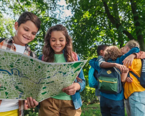 Deux enfants souriants regardent une carte dans un parc, d'autres enfants s'enlacent en arrière-plan.