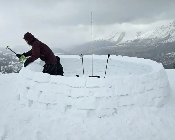 Person construisant un igloo en blocs de neige, avec des bâtons de ski plantés.