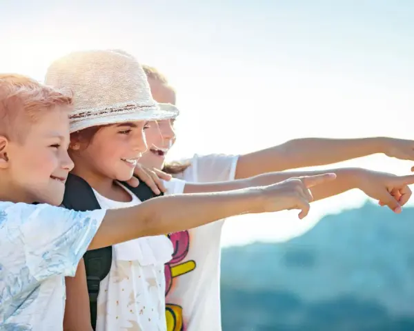Trois enfants souriants, portant des sacs à dos, pointent du doigt vers la droite, sous un ciel ensoleillé.