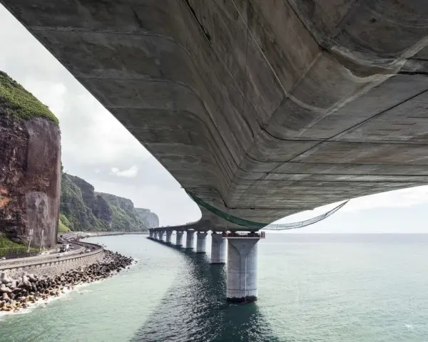 Vue sous un pont en béton, avec la mer, des falaises verdoyantes et une route côtière.