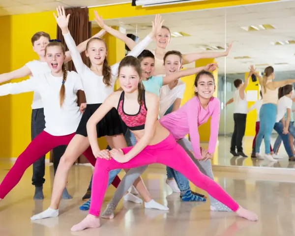 Une joyeuse colonie de danse, composée d'enfants et d'un professeur, s'exerce avec enthousiasme dans un studio lumineux aux murs jaunes.