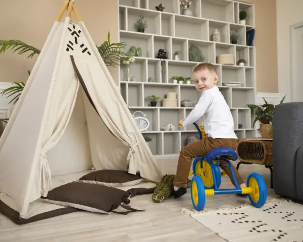 Un petit garçon sur un tricycle bleu et jaune s'amuse dans un espace de jeux, près d'une tente tipi et d'un canapé. Ce centre de vacances et de loisirs est idéal pour les enfants.
