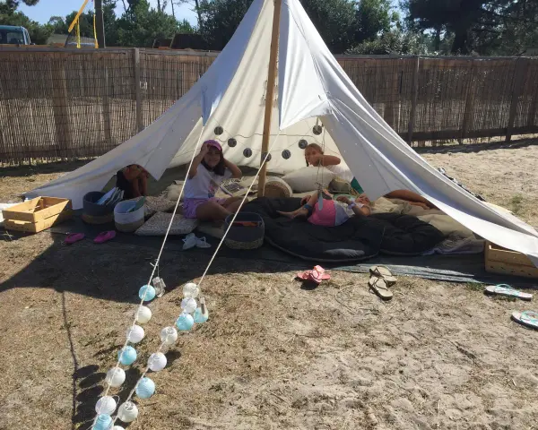 Enfants profitant d'une colonie de vacances camping dans une tente tipi, entourés de coussins et de guirlandes lumineuses.