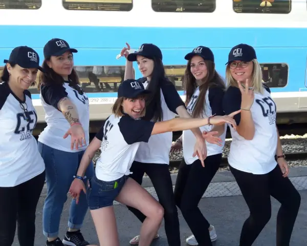 Groupe de jeunes femmes souriantes portant des casquettes et t-shirts "CEI 1947" devant un train.