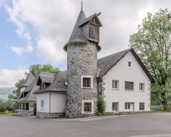 Bâtiment blanc avec une tour ronde en pierre et un toit en ardoise, entouré d'arbres et de verdure.