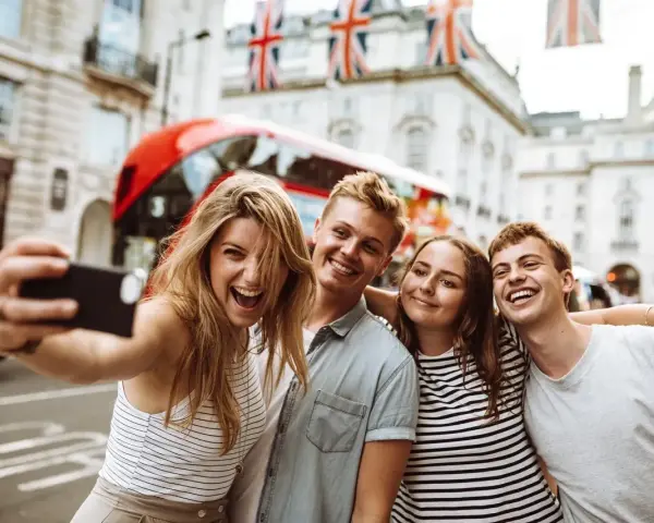 Quatre jeunes souriants prennent un selfie à Londres, avec un bus rouge et des drapeaux britanniques en arrière-plan.