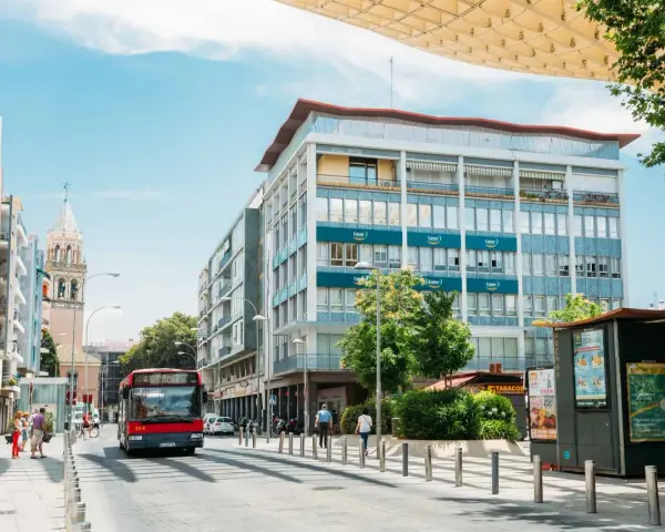 Un bus rouge circule dans une rue bordée d'immeubles et d'un clocher, sous un ciel bleu.