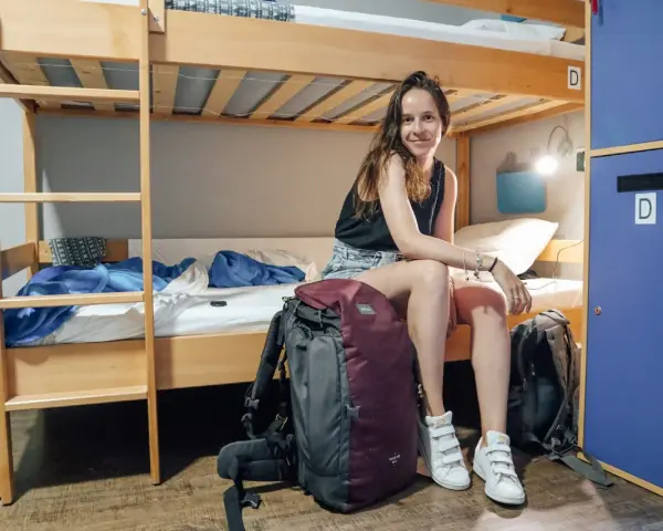 Une jeune femme souriante est assise sur un lit superposé dans un dortoir de colonie de vacances, ses sacs à dos à ses côtés.