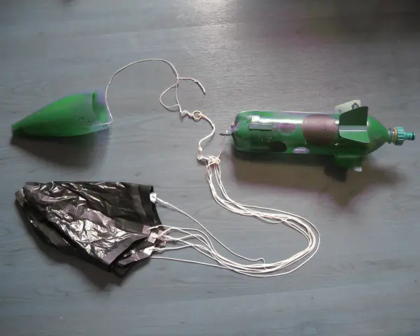 Fusée à eau verte avec cône, parachute noir et fils blancs sur fond gris.