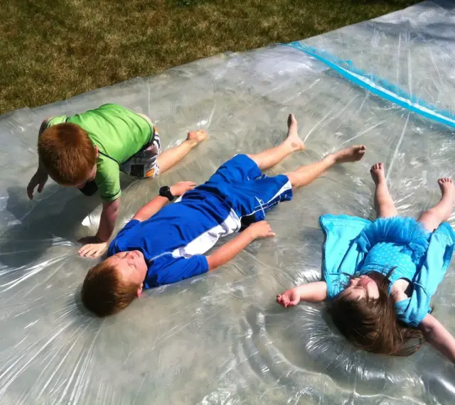 Trois enfants s'amusent sur un jeu d'eau pour kermesse, glissant sur une bâche mouillée sous le soleil.