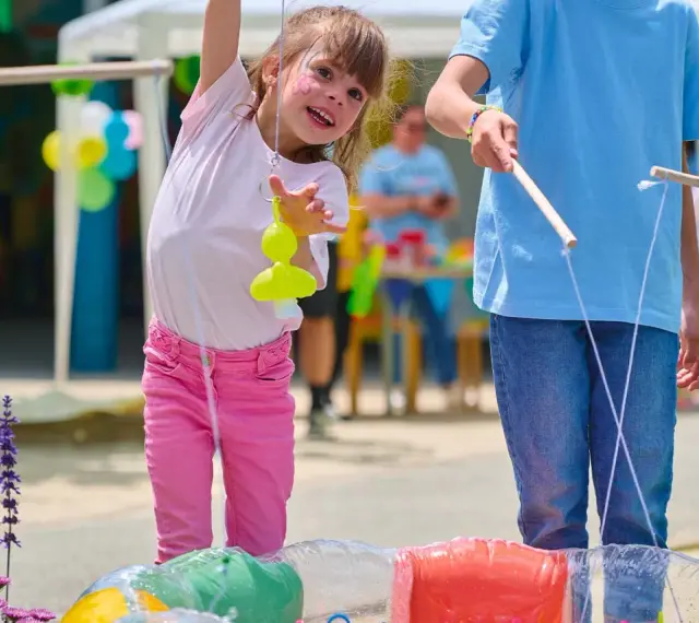 Une petite fille joyeuse participe à des jeux de kermesse pour tout petit, essayant d'attraper un jouet jaune avec une canne à pêche improvisée.