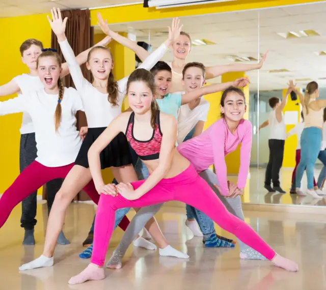 Une joyeuse colonie de danse, composée d'enfants et d'un professeur, s'exerce avec enthousiasme dans un studio lumineux aux murs jaunes.