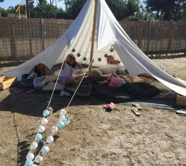 Enfants profitant d'une colonie de vacances camping dans une tente tipi, entourés de coussins et de guirlandes lumineuses.