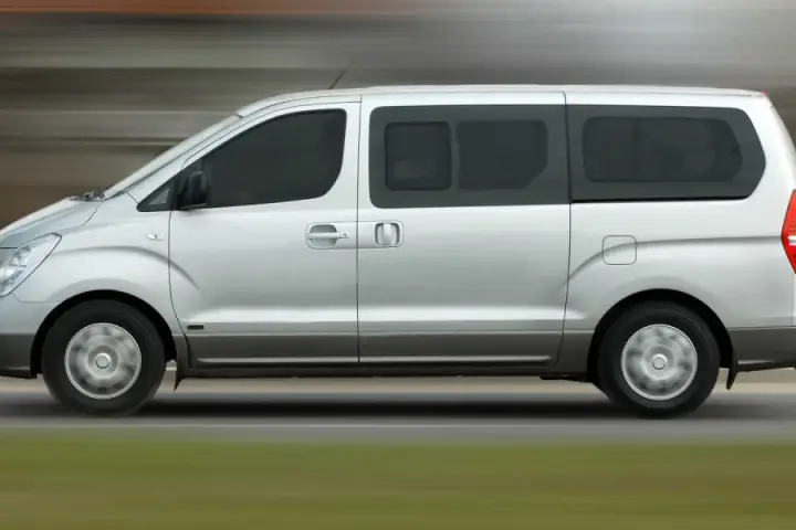 Un mini bus argenté roule sur une route. Ce véhicule spacieux est idéal pour les familles ou les groupes, et peut être conduit avec un permis B.