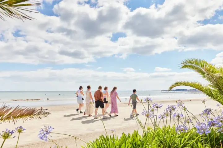 Famille profitant des vacances scolaires en Bretagne, marchant sur la promenade face à la mer, avec des palmiers et des fleurs bleues.