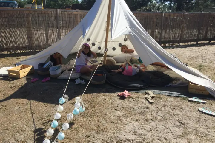 Enfants profitant d'une colonie de vacances camping dans une tente tipi, entourés de coussins et de guirlandes lumineuses.