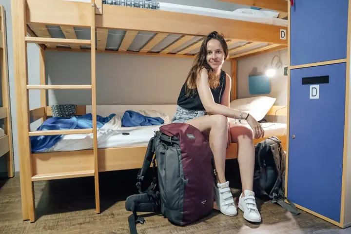 Une jeune femme souriante est assise sur un lit superposé dans un dortoir de colonie de vacances, ses sacs à dos à ses côtés.
