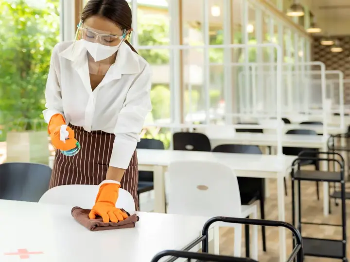 Employée portant masque et visière nettoie une table, respectant la réglementation hygiène cantine scolaire. Des séparateurs sont installés.
