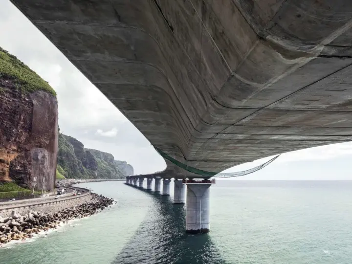 Vue sous un pont en béton, avec la mer, des falaises verdoyantes et une route côtière.