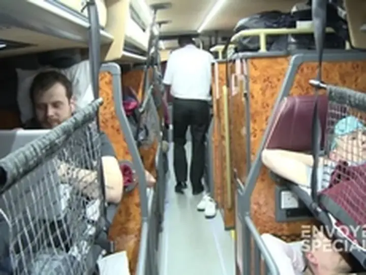 Intérieur d'un bus avec des couchettes. Un homme en blanc marche dans l'allée, tandis que des passagers se reposent dans leurs couchettes.