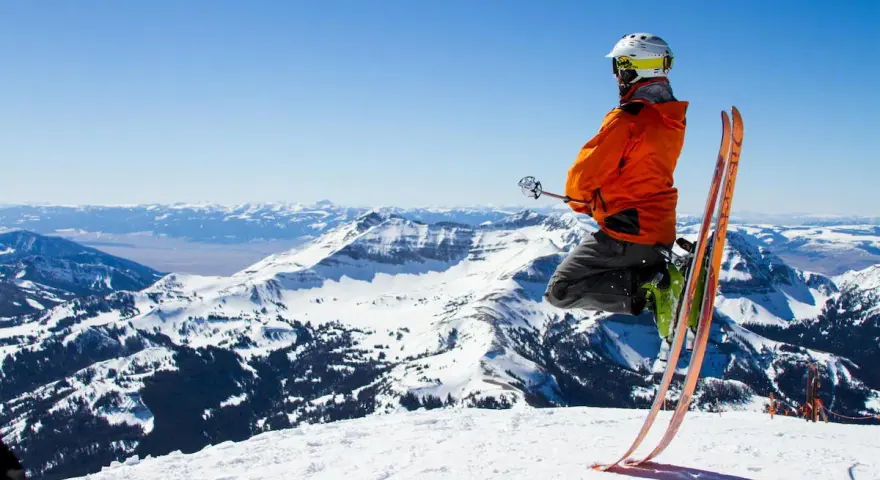 Un skieur en l'air, prêt à dévaler la pente. Il porte un casque et une veste orange vif, rappelant les règles pour skier en toute sécurité. Le paysage montagneux enneigé est spectaculaire.