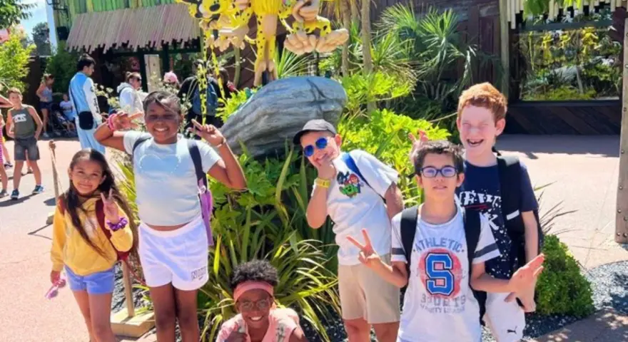 Des enfants joyeux font des signes de paix devant une attraction, un avis "supernova juniors" est visible.