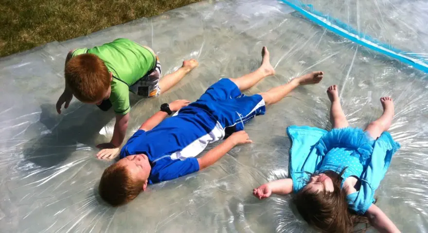 Trois enfants s'amusent sur un jeu d'eau pour kermesse, glissant sur une bâche mouillée sous le soleil.