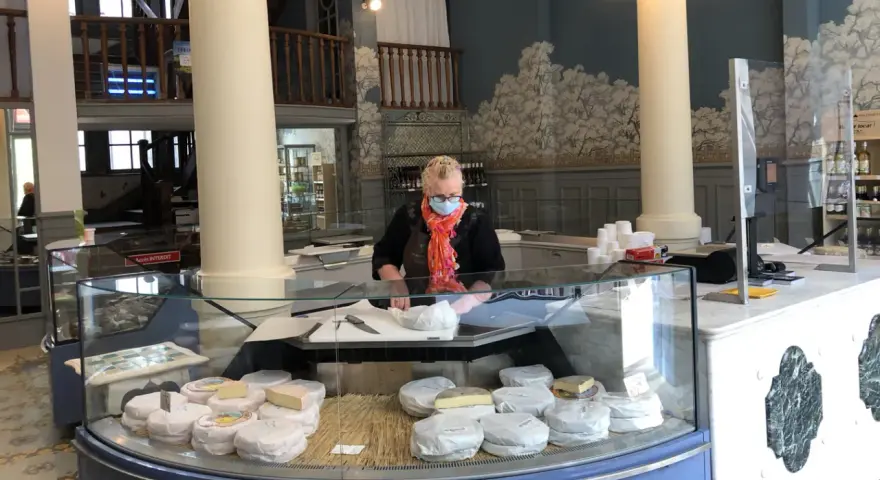 Une fromagère masquée prépare des fromages dans une vitrine réfrigérée.
