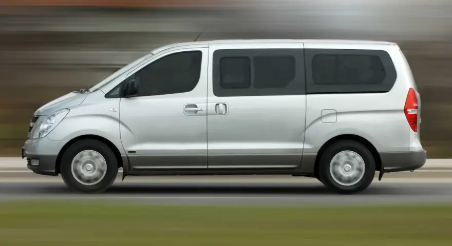 Un mini bus argenté roule sur une route. Ce véhicule spacieux est idéal pour les familles ou les groupes, et peut être conduit avec un permis B.