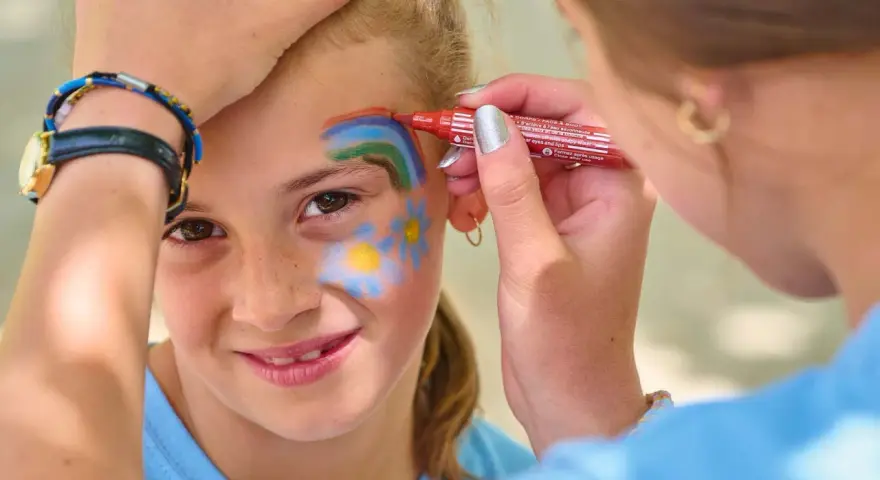 Une petite fille souriante se fait maquiller sur la joue un arc-en-ciel et des fleurs, au stand maquillage d'une kermesse.