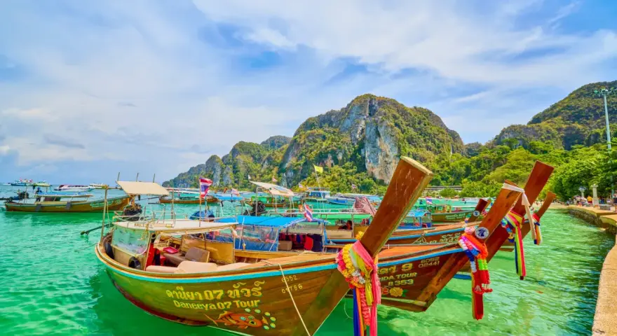 Bateaux traditionnels thaïlandais amarrés dans une eau turquoise, avec des montagnes verdoyantes en arrière-plan.