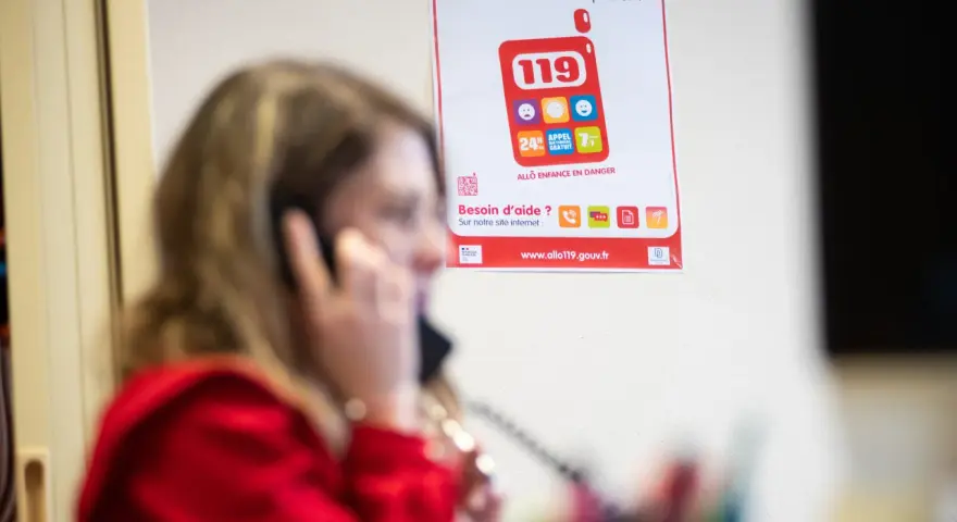 Une femme au téléphone, devant une affiche expliquant le crip signification du numéro 119 pour les enfants en danger.
