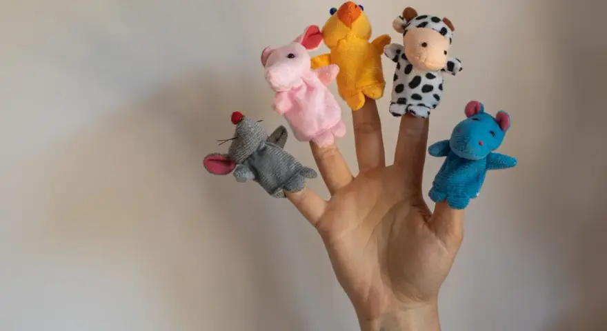 Main d'une personne avec cinq marionnettes à doigts en feutrine : souris grise, lapin rose, canard jaune, vache noire et blanche, hippocampe bleu.
