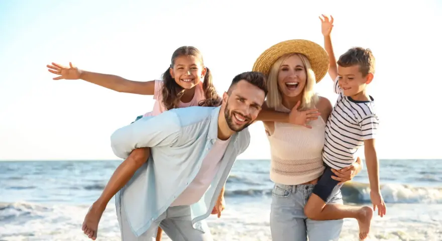 Famille souriante sur la plage, père portant sa fille sur le dos, mère tenant son fils.