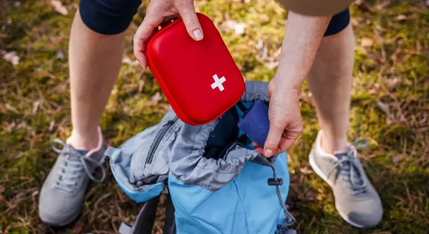 Une personne prépare son sac à dos pour une randonnée, rangeant une trousse à pharmacie enfant rouge avec une croix blanche.