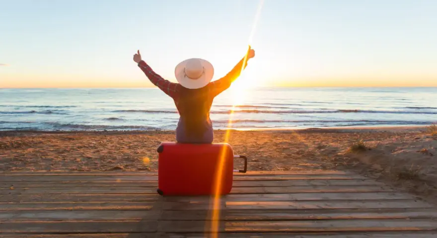 Personne assise sur une valise rouge sur une plage au coucher du soleil, bras levés en signe de victoire.